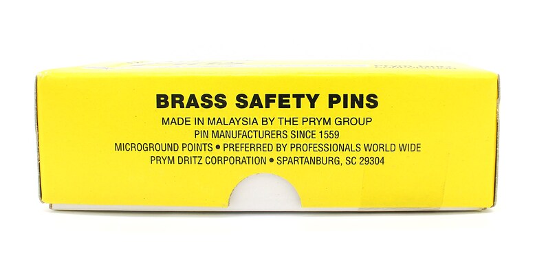 May include: A yellow box with black text that reads "Brass Safety Pins" and "Made in Malaysia by the Prym Group". The box also states "Pin Manufacturers Since 1559", "Microground Points", "Preferred by Professionals World Wide", "Prym Dritz Corporation", and "Spartanburg, SC 29304".