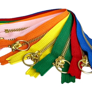 Könnte beinhalten: Ein Satz von acht bunten Reißverschlussbändern mit goldenen Metall-Schiebern. Die Bänder sind in einem Regenbogenmuster angeordnet, mit rosa, orange, gelb, grün, blau, rot und zwei Gelbtönen.