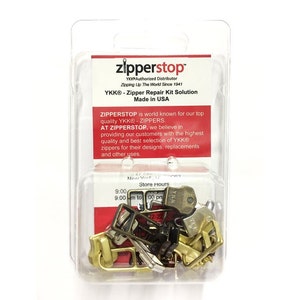 May include: A clear plastic package containing a zipper repair kit solution. The kit includes a variety of metal zipper sliders, pulls, and stops in gold and black. The package is labeled "zipperstop" and "YKK® - Zipper Repair Kit Solution Made in USA".
