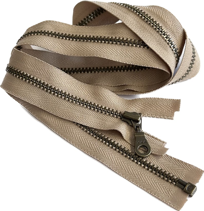 May include: A beige zipper with a brass finish. The zipper is closed and has a pull tab with the brand name "YKK" on it.