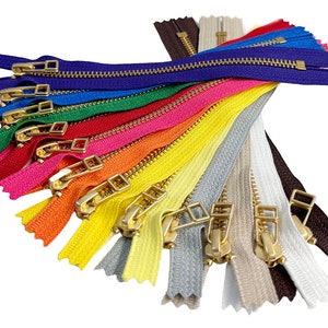 May include: A collection of colorful zippers with gold-colored metal pulls. The zippers are arranged in a fan shape, showcasing a variety of colors including blue, green, red, pink, orange, yellow, gray, and brown. The image is set against a white background.