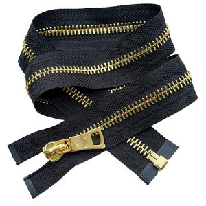 May include: A black zipper featuring metallic gold-toned teeth and a matching gold-toned metal slider pull. The zipper tape is made of a woven black fabric, and the teeth are arranged in a continuous row along the length of the fastener.