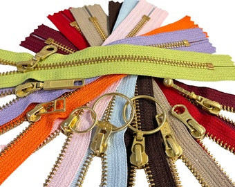 May include: A collection of colorful zippers in various shades, including green, orange, purple, and pink. Each zipper has a brass-colored pull and teeth. The zippers are arranged in a fan shape against a white background.