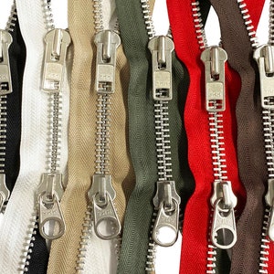 Op de afbeelding: Een set van acht metalen ritsen in verschillende kleuren, waaronder zwart, wit, beige, groen, rood en bruin. Elke rits heeft een zilveren treklip met het YKK-logo.
