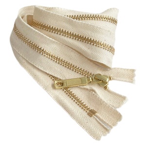 May include: A close-up of a beige zipper with a gold metal slider and pull tab. The zipper is made of cotton and has a herringbone pattern.