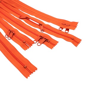 May include: A close-up of a pile of orange zipper closures. The zippers are arranged in a fan shape, with the teeth facing up. The zippers are made of a durable nylon material and have a smooth, easy-to-use slider.