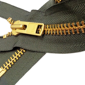 May include: Close-up of a dark green zipper with gold-colored teeth and a matching pull tab. The zipper is partially open, revealing the interlocking teeth. The YKK brand is visible on the pull tab.