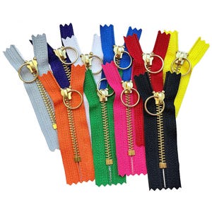 May include: A collection of colorful zippers with gold-tone metal pulls. The zippers are in various colors, including orange, green, pink, red, black, and yellow. Each zipper has a ring pull.