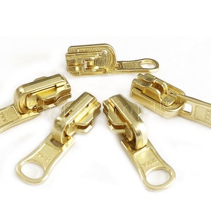 May include: Five gold-coloured zipper pulls with the brand name YKK printed on each. The zipper pulls are arranged in a circle on a white background.
