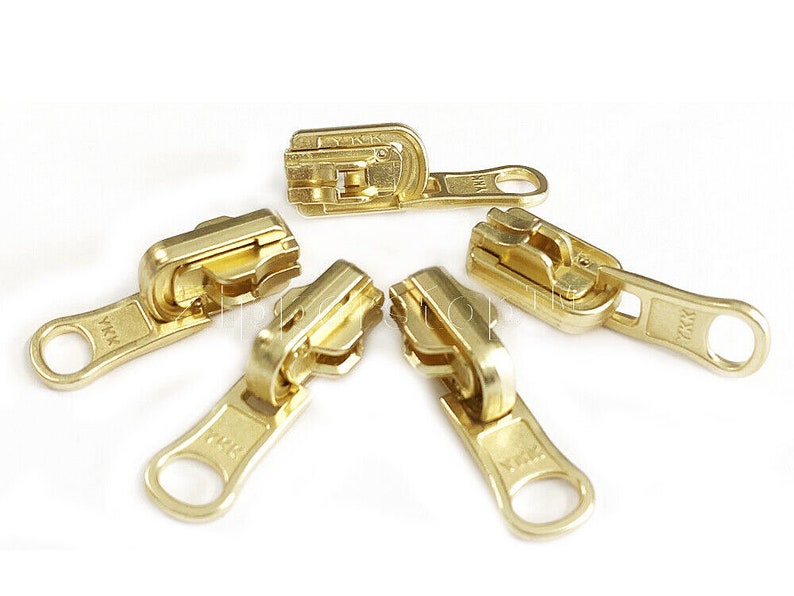 May include: Five gold-coloured zipper pulls with the brand name YKK printed on each. The zipper pulls are arranged in a circle on a white background.