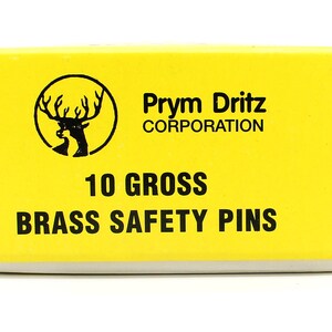 May include: A yellow box with black text that reads "Prym Dritz Corporation", "10 Gross", and "Brass Safety Pins". A black and white image of a deer head with antlers is in the top left corner of the box.