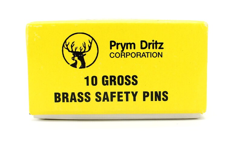 May include: A yellow box with black text that reads "Prym Dritz Corporation", "10 Gross", and "Brass Safety Pins". A black and white image of a deer head with antlers is in the top left corner of the box.
