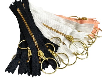 May include: A collection of zippers in various colors, including black, white, and peach. Each zipper features a gold-colored metal pull and a circular ring attached to the slider. The zippers are arranged in a neat stack, ready for use in sewing or crafting projects.