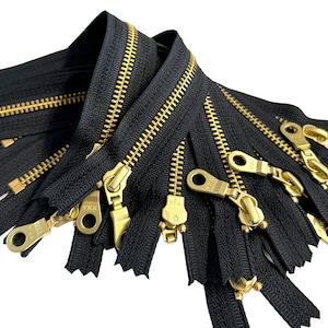 Könnte beinhalten: Nahaufnahme eines Stapels schwarzer Reißverschlüsse mit goldenen Metallzähnen und Schiebern. Die Reißverschlüsse sind mit "YKK" gekennzeichnet und eignen sich ideal für Nähprojekte.