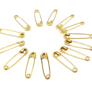 May include: A set of 12 gold safety pins arranged in a circle on a white background.