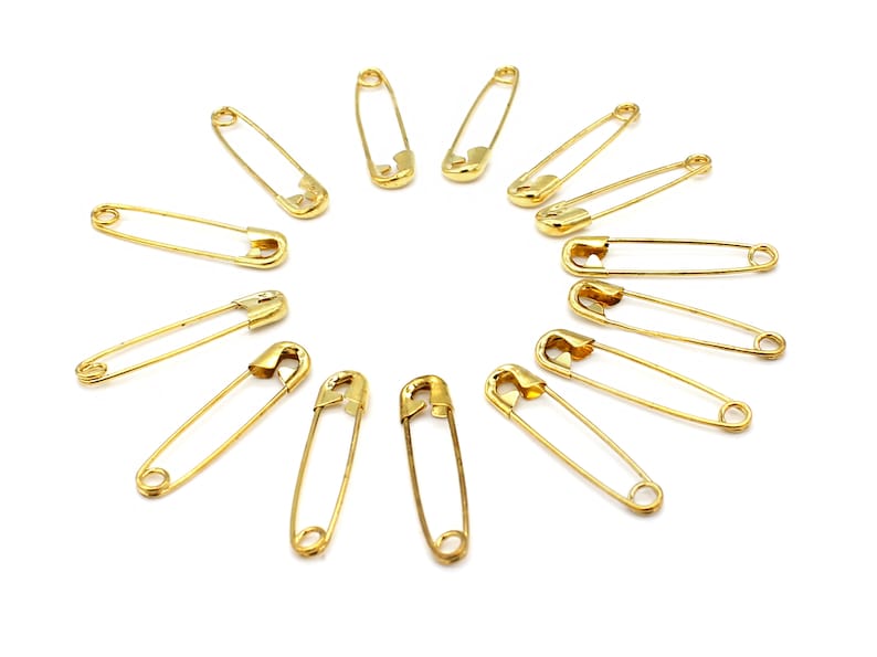 May include: A set of 12 gold safety pins arranged in a circle on a white background.