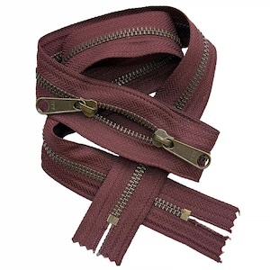 May include: A coiled burgundy zipper featuring antique brass-colored metal teeth and two matching metal sliders. The fabric tape has a visible herringbone weave pattern, and the YKK brand name is embossed on the metal pull tabs of the sliders.