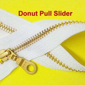 Pode incluir: Um z&iacute;per branco com um cursor de metal dourado. O cursor tem um anel em forma de rosquinha e o texto "YKK" &eacute; vis&iacute;vel no cursor. O z&iacute;per est&aacute; sobre um fundo amarelo.