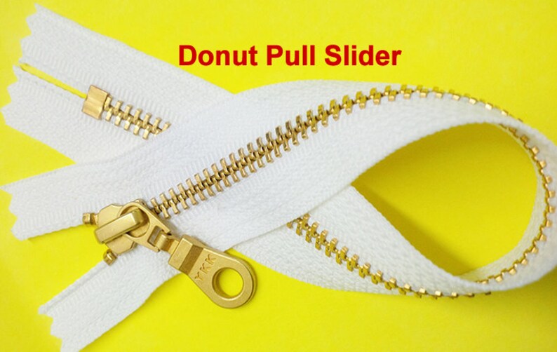 Pode incluir: Um z&iacute;per branco com um cursor de metal dourado. O cursor tem um anel em forma de rosquinha e o texto "YKK" &eacute; vis&iacute;vel no cursor. O z&iacute;per est&aacute; sobre um fundo amarelo.
