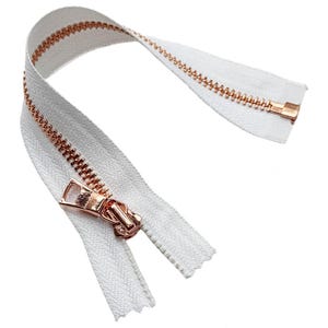 May include: A white zipper featuring metallic rose gold teeth and a matching rose gold pull tab. The zipper tape is made of a woven white fabric, and the metal teeth are arranged in a continuous row along the length of the fastener.