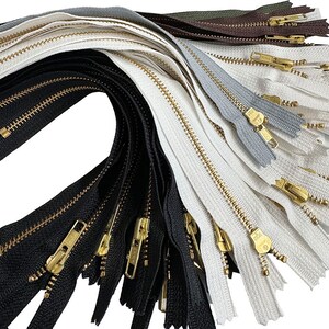 May include: A collection of various colored zippers with gold-toned metal pulls. The zippers are in shades of black, white, gray, and brown. The image is a close-up shot, highlighting the details of the zipper teeth and pulls.
