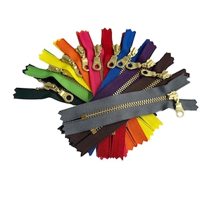May include: A selection of colourful zippers with gold-coloured pulls, arranged in a fan shape. The zippers are in various colours, including green, yellow, orange, pink, red, blue, purple, brown, and grey. The image is on a white background.