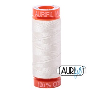 May include: A spool of white Aurifil thread with the text "Aurifil Made in USA" on the top and "100% Cotton" on the bottom.