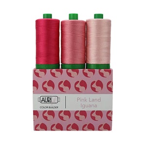 May include: Three spools of pink thread in a color gradient from dark pink to light pink. The spools are in a box with a pink and white pattern of stylized iguanas. The box has the text "Pink Land Iguana" and "AURIfil Color Builder" on it.