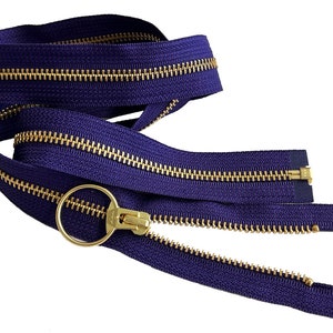 Puede incluir: Primer plano de una cremallera morada con un deslizador y una lengüeta de metal dorado. La cremallera está dispuesta en un bucle sobre un fondo blanco.