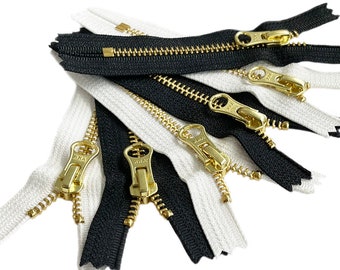 May include: Close-up of several zippers in black and white with gold-colored metal pulls. The zippers are arranged in a pile, showcasing their teeth and pull tabs. The image is well-lit, highlighting the details of the sewing notions.