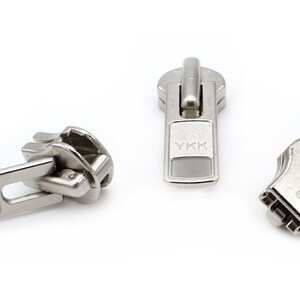 May include: Three silver metal zipper pulls with the YKK logo. The zipper pulls are in different styles.