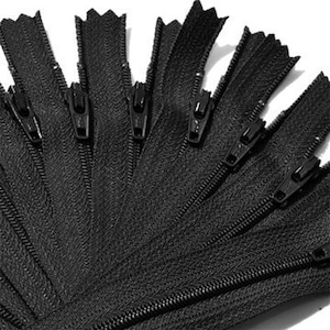 May include: Close-up of several black zippers fanned out against a white background. Each zipper has a black pull tab and teeth. The zippers are made of a durable material and are suitable for various sewing projects.