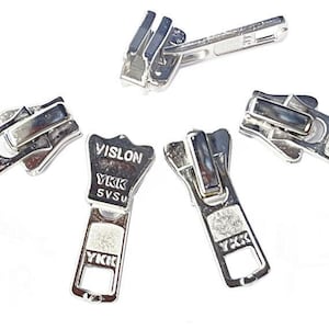 May include: A collection of silver-colored zipper pulls, some with the "VISLON YKK SVSU" brand name. The zipper pulls are made of metal and are designed for use in clothing or other fabric items. The image is taken against a white background.