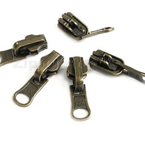 May include: Five antique gold zipper pulls with a YKK logo. The zipper pulls have a loop on one end and a slider on the other end.
