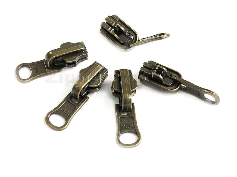 May include: Five antique gold zipper pulls with a YKK logo. The zipper pulls have a loop on one end and a slider on the other end.