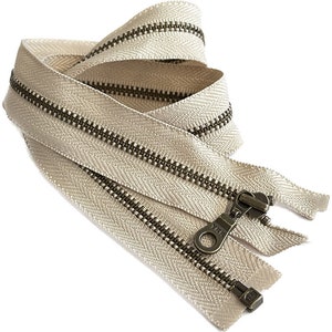 Pode incluir: Um zíper bege com dentes de latão antigo e um cursor. O zíper está dobrado ao meio e o cursor é visível.