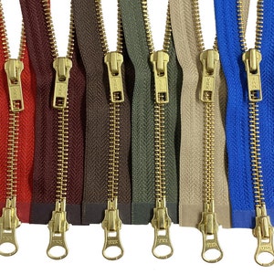 Puede incluir: Una colección de cremalleras metálicas de colores con tiradores dorados. Las cremalleras son de varios colores, incluyendo rojo, burdeos, marrón, verde oliva, beige y azul. Cada cremallera está cerrada y se muestra verticalmente.