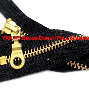 May include: A black zipper with a gold YKK #5 brass donut pull slider. The zipper is closed and the slider is in the center.