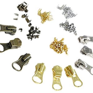 以下が含まれることがあります： ゴールド、シルバー、ブロンズ仕上げのYKKジッパーのプルタブの詰め合わせ。プルタブは白い背景に円形に配置されています。各プルタブはスタイルとサイズが異なります。