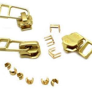 K&ouml;nnte beinhalten: Eine Sammlung goldfarbener Rei&szlig;verschlusskomponenten, darunter Zipper, Schieber und verschiedene Befestigungselemente. Die Komponenten sind auf wei&szlig;em Hintergrund angeordnet und zeigen ihr Design und ihre Konstruktion. Ideal f&uuml;r N&auml;h- und Bastelprojekte.