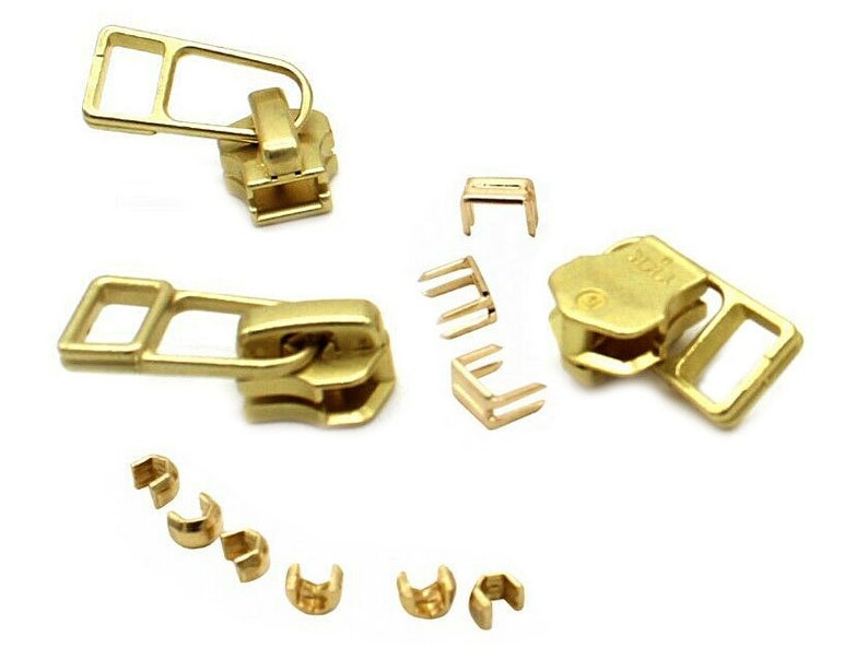 K&ouml;nnte beinhalten: Eine Sammlung goldfarbener Rei&szlig;verschlusskomponenten, darunter Zipper, Schieber und verschiedene Befestigungselemente. Die Komponenten sind auf wei&szlig;em Hintergrund angeordnet und zeigen ihr Design und ihre Konstruktion. Ideal f&uuml;r N&auml;h- und Bastelprojekte.