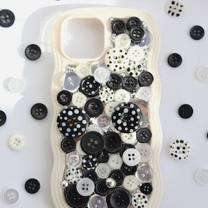 Op de afbeelding: Een crèmekleurig telefoonhoesje versierd met een dichte collage van diverse zwarte, witte en doorzichtige knopen van verschillende groottes en patronen. De knopen zijn over de achterkant van het hoesje gelaagd, met stippen, effen kleuren en texturen.