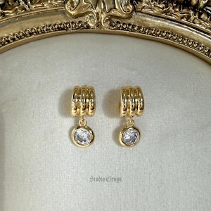 Puede incluir: Un par de pendientes de aro dorados con un colgante de piedra preciosa redonda y transparente. Los pendientes tienen un diseño acanalado en el aro y se exhiben sobre una superficie de color claro. La marca "Studio Elevyn" es visible.