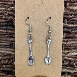 May include: A pair of silver earrings featuring a fork and a spoon charm. The earrings are hanging from a brown card.