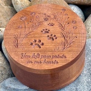 May include: A round, wooden keepsake box with a dark brown finish. The lid is engraved with paw prints, floral accents, and the text "You left paw prints on our hearts". The box is sitting on a bed of rocks.