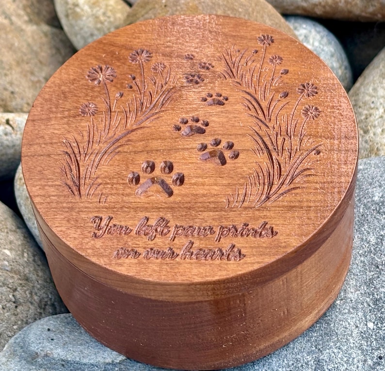 May include: A round, wooden keepsake box with a dark brown finish. The lid is engraved with paw prints, floral accents, and the text "You left paw prints on our hearts". The box is sitting on a bed of rocks.