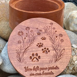 May include: A wooden pet urn with a lid. The lid is engraved with paw prints, flowers, and the text "You left paw prints on our hearts." The urn and lid are a reddish-brown color.