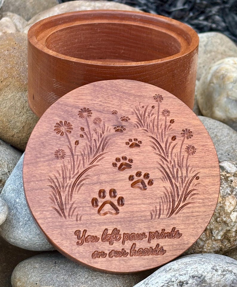 May include: A wooden pet urn with a lid. The lid is engraved with paw prints, flowers, and the text "You left paw prints on our hearts." The urn and lid are a reddish-brown color.