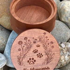 May include: A round, wooden keepsake box with a lid. The lid is engraved with paw prints, flowers, and the text "You left paw prints on our hearts." The box is made of reddish-brown wood.