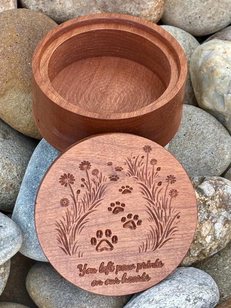 May include: A round, wooden keepsake box with a lid. The lid is engraved with paw prints, flowers, and the text "You left paw prints on our hearts." The box is made of reddish-brown wood.
